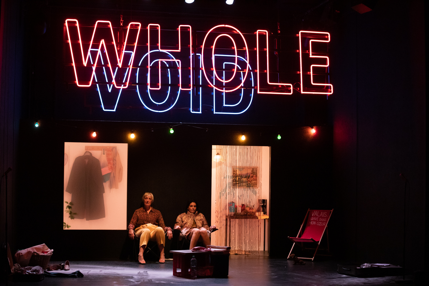 Photo représentant un moment de la pièce. Deux comédiennes sont assises en fond de scène sous des mots écrits en néon rouge et bleu. Elles attendent. Photo prise par Rebecca Bowring