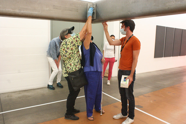 Cette photo représente des personnes aveugles et malvoyantes. Ces personnes participent à une visite tactile au MAMCO, et sont en train de découvrir une œuvre en béton, fixée au plafond.