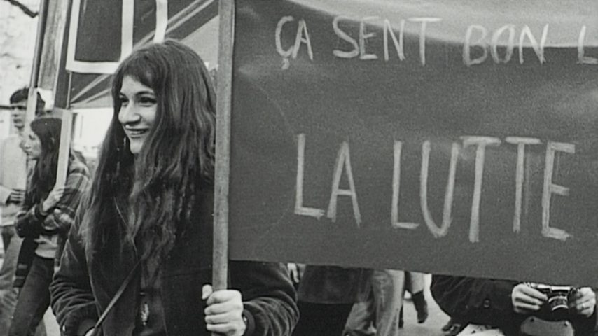 L'image est en noir et blanc. On y voit une jeune femme au premier plan qui manifeste avec d'autres jeunes. Elle tient une banderole sur laquelle on lit "La Lutte".