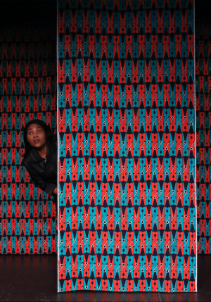 Une femme se trouve derrière un panneau aux motifs colorés rouges et bleus. Elle apparait sur la gauche, pour regarder le public.