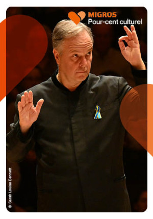 L'image montre un chef d'orchestre. Il est de trois-quart. Sa main gauche est levée dans un mouvement de direction d'orchestre. Il est habillé en noir et regard droit devant lui.