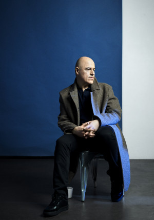 L'image montre un homme assis. Il est chauve, porte un manteau et regarde sur sa gauche. Derrière lui, le mur est bleu et blanc.
