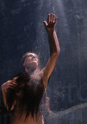 La photo montre une femme avec de longs cheveux foncés derrière un plexiglas. Elle regarde en l'air et tend la main en l'air aussi, collée contre le plexiglas. La lumière vient d'en haut, elle illumine la femme.