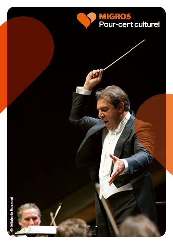 La photo montre un chef d'orchestre en train de diriger un orchestre. Il a dans sa main droite une baguette qu'il lève dans un mouvement vif. Son regard est posé sur les musiciens devant lui.