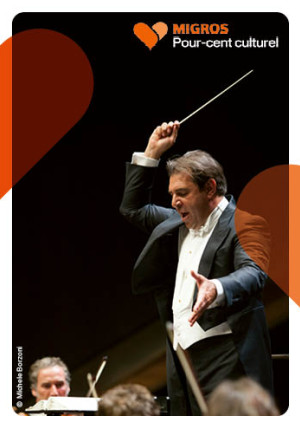 La photo montre un chef d'orchestre en train de diriger un orchestre. Il a dans sa main droite une baguette qu'il lève dans un mouvement vif. Son regard est posé sur les musiciens devant lui.